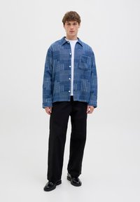 Modrá károvaná košeľa so zapínaním na gombíky a jedným náprsným vreckom, v kombinácii s čiernymi nohavicami a lesklými čiernymi topánkami na bielom pozadí.