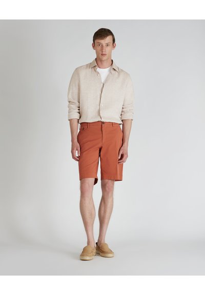 Pantalones cortos color óxido hechos de una tela con textura suave, combinados con una camisa de lino de botones de color claro. Llevados con zapatos de lona beige sin cordones.