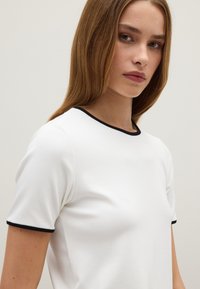 Maglietta bianca a maniche corte con colletto a coste nere. Tessuto liscio con un design slim, caratterizzato da cuciture minime.