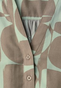 Gros plan sur un tissu beige et vert clair à motifs avec un col en V et deux boutons blanc cassé portant l'inscription "Street One".