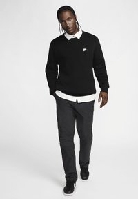 Μαύρο πουλόβερ πλεκτό με v-λαιμόκοψη και λευκό λογότυπο Nike, τοποθετημένο πάνω από λευκή μπλούζα με γιακά; συνδυασμένο με σκούρα, υφασμάτινα παντελόνια και μαύρα παπούτσια.