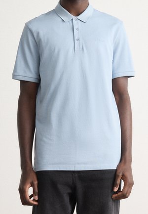 Mand iført lyseblå kortærmet poloshirt med tre knapper, kombineret med sorte bukser, stående foran en ensfarvet baggrund.