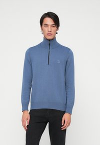 Blauwe langarm pullover met een hoge kraag en een ritssluiting aan de voorkant, voorzien van een gestructureerde brei en een logo patch op de borst.
