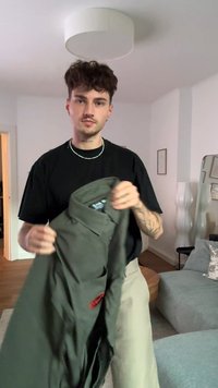 Una camicia verde con bottoni, tenuta da una persona, presenta un colletto, una tasca frontale e dettagli rossi. Il tessuto appare liscio e leggero.