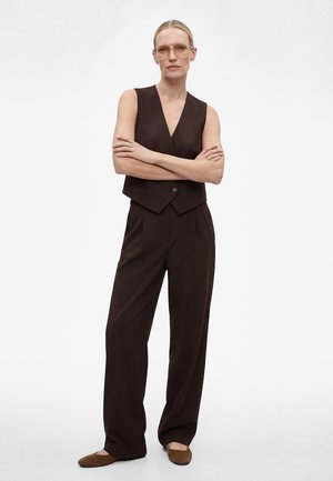 Kobieta stoi ze skrzyżowanymi ramionami, ubrana w brązową kamizelkę bez rękawów, pasujące szerokie spodnie, brązowe baleriny i przezroczyste okulary.