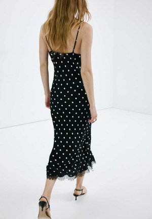 Femme aux longs cheveux marchant dos à nous, vêtue d'une robe midi noire à pois blancs et de sandales à talons noires dans une pièce blanche.