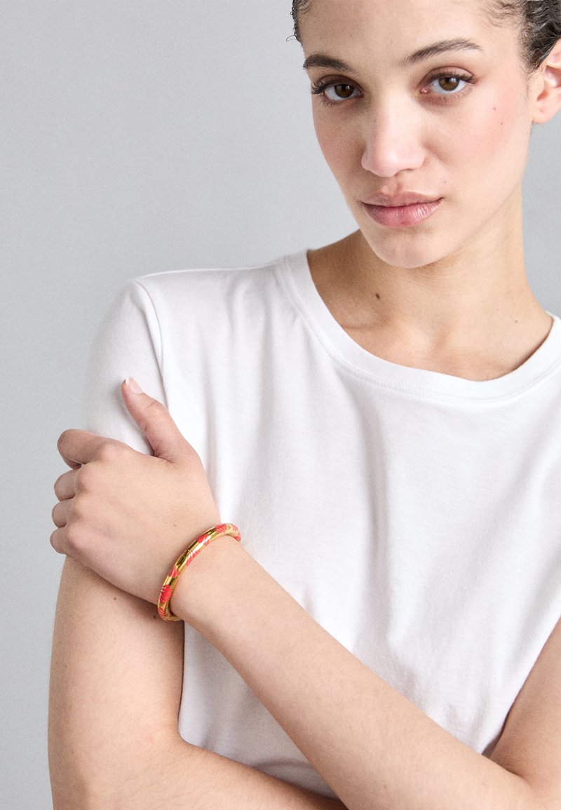 Barevný náramek nošený na zápěstí, s květinovým vzorem na texturovaném pozadí. V pozadí neformální bílé tričko.