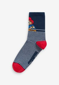 Sininen ja valkoinen raidallinen sukka, jossa on punainen varvas ja kantapää, esittää kuvittettua karhua punaisessa hatussa ja sinisessä takissa sekä "Paddington"-teksti.