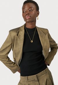 Bruin geruit blazer met notch lapels, gecombineerd met een zwart geribbeld topje. Gouden hangerketting en knoopdetails op de blazer zijn zichtbaar.