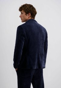 Man in een blauw corduroy pak met handen in de zakken, die zijwaarts kijkt tegen een effen witte achtergrond.