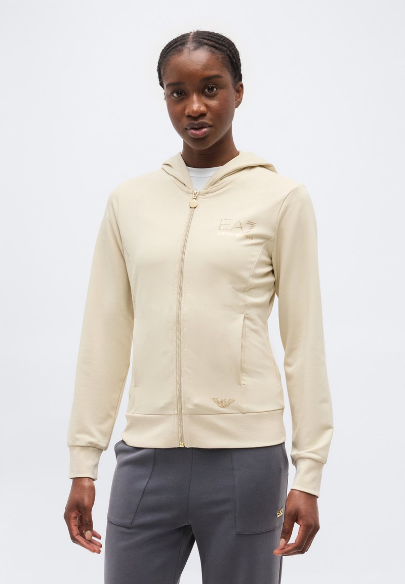 Žena s vrkočmi, oblečená v béžovej mikine s kapucňou na zips a sivých nohaviciach, stojaca pred jednoduchým svetlým pozadím