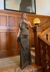 Femme en robe une épaule vert olive brillante avec des détails en dentelle pose sur un escalier en bois tenant une pochette noire sous une lampe allumée.