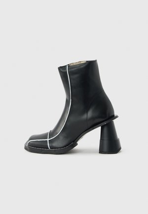 Stivaletto alla caviglia in pelle nera con cuciture bianche, punta quadrata e tacco largo svasato unico, mostrato di profilo su sfondo grigio.