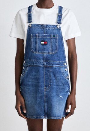 Person iført blå denim overalls med forlomme og "Tommy Jeans" logo, kombineret med en enkel hvid kortærmet skjorte.