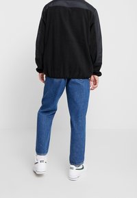 Chaqueta tipo pulóver en negro y gris con tela texturizada, vaqueros azules y zapatillas blancas con detalles en verde. Costuras visibles.