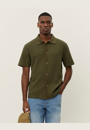 Olive green short-sleeve shirt with a textured, waffle-knit design, button-down front, and a classic collar. Worn with light blue jeans.