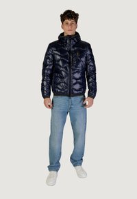 Giacca pesante blu navy con zip frontale e cappuccio, con texture trapuntata e tasca sul petto. Abbinata a jeans azzurri e scarpe bianche.