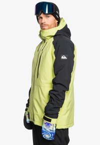 Skridskojacket i klar limegrön och svart, med huva, dragkedjefickor och justerbara ärmslut. I kombination med blå och vita mönstrade handskar.