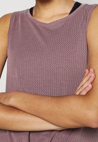 Personne aux bras croisés portant un haut violet texturé sans manches avec un sous-vêtement noir visible au niveau du col.