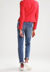 Rød tekstureret blazer med lange ærmer, parret med lyseblå oprullede jeans og hvide sneakers med røde accenter.