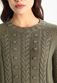 Pull en tricot vert olive avec des motifs en relief, des manches texturées et un col côtelé, agrémenté de boutons à l'avant.