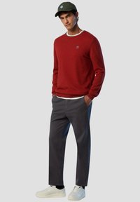 Maglione rosso con scollo rotondo e logo ricamato, abbinato a pantaloni grigio scuro e sneaker bianche. Il modello indossa un cappellino verde.