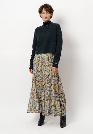 Vrouw in een donzige zwarte trui, bloemenmaxi-rok en zwarte enkellaarsjes tegen een effen witte achtergrond.