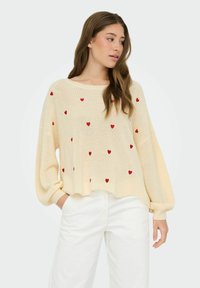 Jeune femme aux longs cheveux portant un pull en maille crème avec des cœurs rouges et un pantalon blanc, debout avec une main dans sa poche.