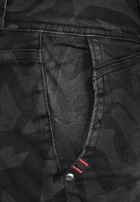 Svarta denimjeans med ett subtilt camouflage-mönster, med en silverknapp och röda stickningar som detaljer på fickan. Strukturerad tyg.