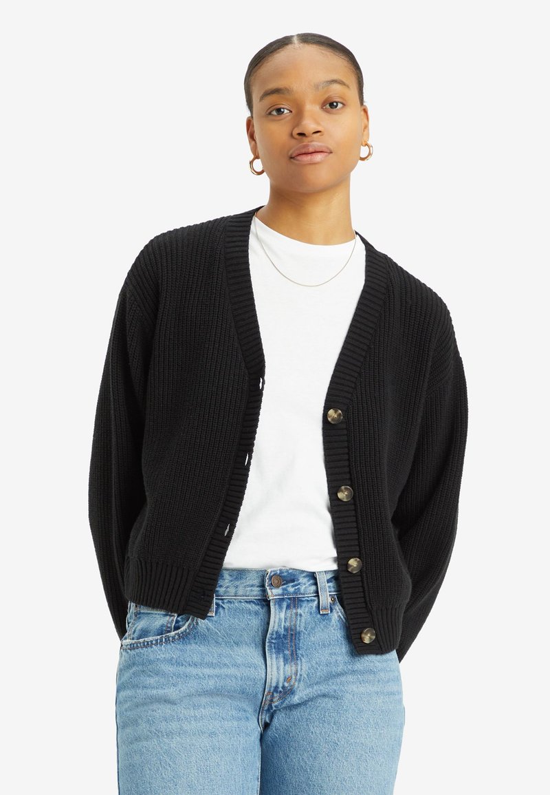 Cardigan noir à côtes avec un col en V, doté de cinq boutons, porté par-dessus un t-shirt blanc, associé à un jean en denim bleu clair.