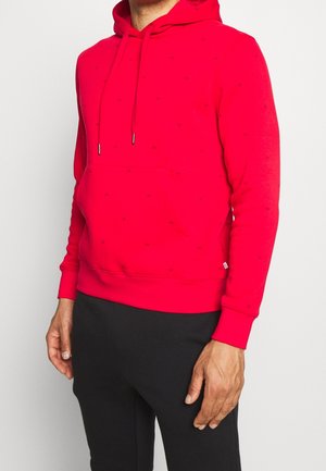 Uomo che indossa una felpa con cappuccio rosso brillante con tasca frontale e pantaloni neri, in piedi contro uno sfondo bianco uniforme.