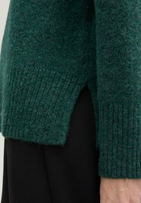 Pulover verde tricotat, cu hem și fante laterale ribbed. Textură moale, cu mâneci lungi și un croi lejer, asortat cu pantaloni negri.