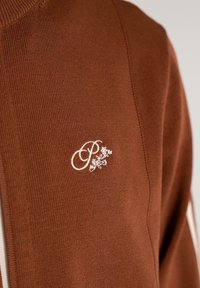 Brun strikket stof med hvid broderet bogstav "P" og små blomsterdesign nær midten.