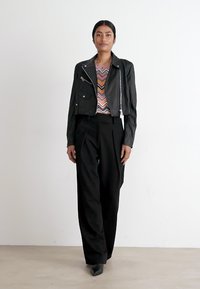 Veste en cuir noir coupée sur un haut à motif zigzag coloré, associée à un pantalon noir taille haute. Le mannequin se tient contre un fond neutre.