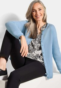 Lachende volwassen vrouw met lang grijs haar zit casual gekleed in een lichtblauwe cardigan, zwarte broek, zwarte sneakers en een gebloemd blouse.