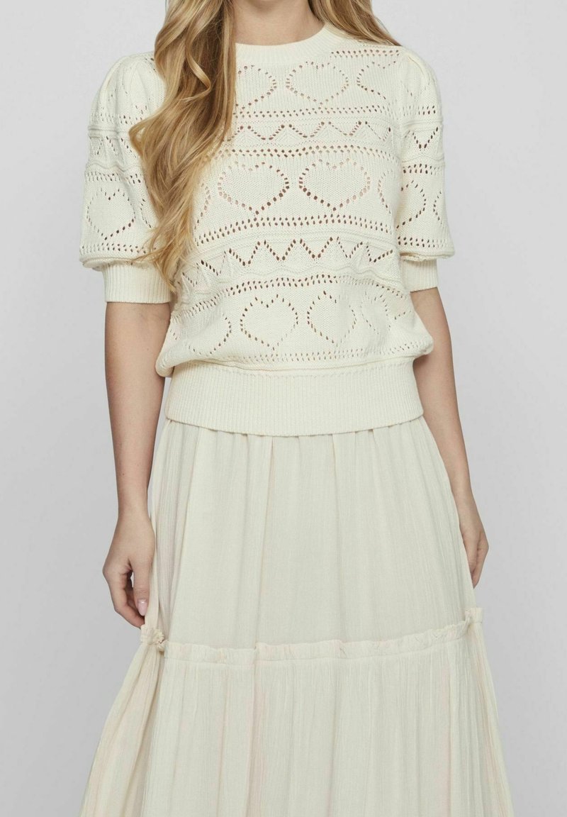Pletený krémový svetr s motivy srdce a cik-cak, krátkými puff rukávy a žebrovým lemováním. V kombinaci s lehkou, světlou maxi sukni.
