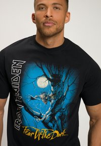 Černé bavlněné tričko s grafickým potiskem monstra na stromě proti modré noční obloze, s texty "Iron Maiden" a "Fear Of The Dark".