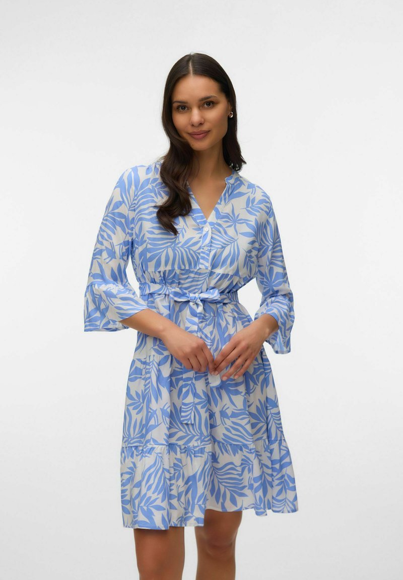 Femme aux longs cheveux foncés portant une robe blanche jusqu'au genou avec des motifs de feuilles bleues, devant boutonné, manches trois-quarts et ceinture à la taille nouée.