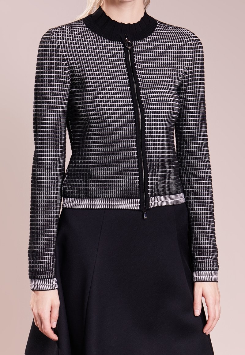 Black and white patterned jacket with a zip closure, ribbed collar, and cuffs. Textured fabric with a fitted silhouette, paired with a black skirt.