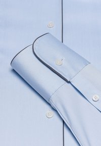 Helles blaues Hemd mit glatter Textur, versehen mit einem kontrastierenden dunklen Saum entlang des Kragens und der Manschetten sowie weißen Knöpfen.
