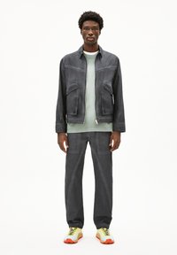 Grauer Reißverschluss-Jacke mit zwei Fronttaschen, kombiniert mit einem hellgrünen Shirt, grauen Hosen und mehrfarbigen Sneakers mit gelben und orangefarbenen Akzenten.