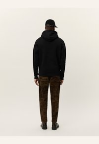 Svart hettegenser med snor, kombinert med brune teksturerte bukser med mønster. Modellen står med ryggen til, iført en svart caps.