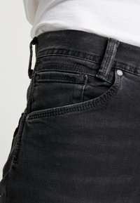 Jeans de mezclilla negra con un diseño ajustado, que cuentan con bolsillos laterales y traseros, costuras visibles y un botón de metal en la cinturilla.