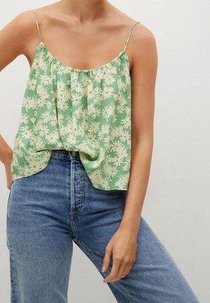 Kobieta ubrana w zieloną, kwiatową bluzkę na cienkich ramiączkach, zestawioną z wysokim stanem niebieskich dżinsów, stojąca z jedną ręką na biodrze.