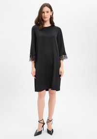 Zwarte satijnen jurk met driekwart mouw met wijde klokmouwen en een ingewikkelde kanten rand. Het ontwerp is losvallend en eindigt boven de knieën.