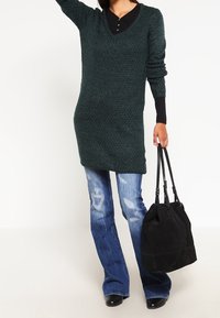 Green knit tunic with a v-neck and long sleeves, paired with faded blue flared jeans. Black handbag with dual handles is held in one hand.