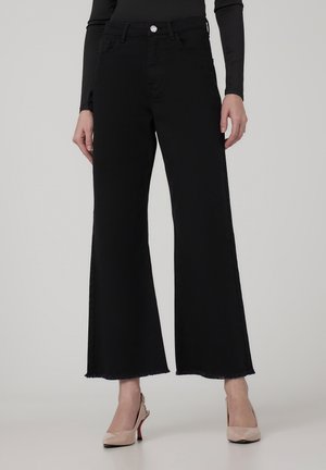 Czarne spodnie dzwony z postrzępionym dołem, wysokim stanem i srebrnym detalem na guzik. Noszone z kremowymi obcasami. Gładka tekstura denimu.