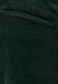 Donkergroene corduroy stof, met een zachte textuur en diagonale ribbels. Bevat een zakdetail dat wordt benadrukt door subtiele stiksels.