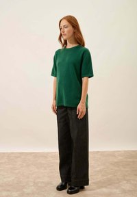 Zielona krótko rękawowa bluzka z mieszanki wełny, zestawiona z ciemnoniebieskimi spodniami wysokiej talii i czarnymi, masywnymi butami na obcasie, stojąca na jasnej powierzchni.