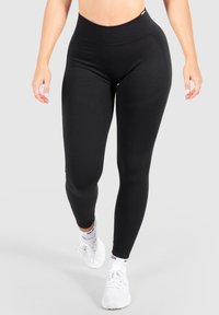 Leggings neri a vita alta realizzati in tessuto elasticizzato, caratterizzati da una texture liscia e da un design aderente, abbinati a scarpe da ginnastica bianche.